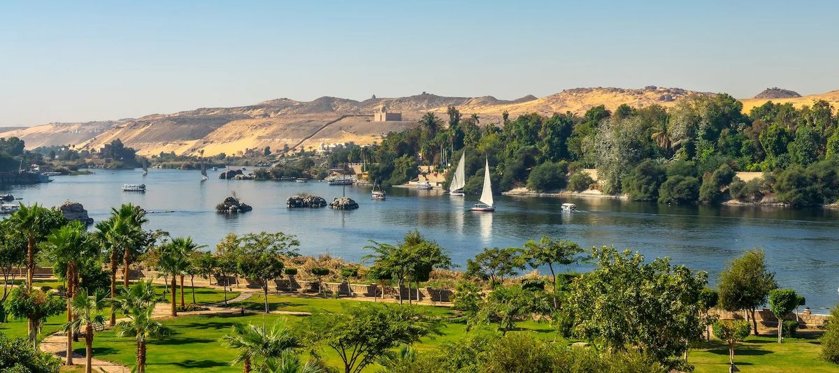 Weiße Segelschiffe gleiten auf dem Nil vor grünen Ufern, während sich im Hintergrund Wüstenberge erheben