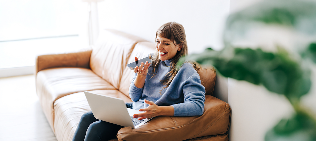 Frau sitzt auf Sofa mit Laptop auf dem Schoß und Telefon in der Hand